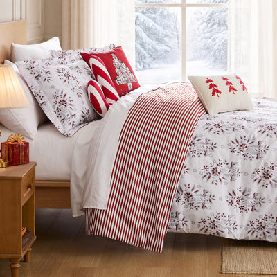 Merry Manor Christmas comforter set styled on a bed with a white ground red and gray floral, poinsettia, and reindeer print, red and white ticking stripe reverse folded back, matching print shams, a red Happy Holidays gift-tree pillow, a candy cane bolster, and a cream red-tree lumbar pillow, beside a lamp-lit nightstand with red gifts and a snowy window view.