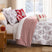 Merry Manor Christmas comforter set styled on a bed with a white ground red and gray floral, poinsettia, and reindeer print, red and white ticking stripe reverse folded back, matching print shams, a red Happy Holidays gift-tree pillow, a candy cane bolster, and a cream red-tree lumbar pillow, beside a lamp-lit nightstand with red gifts and a snowy window view.