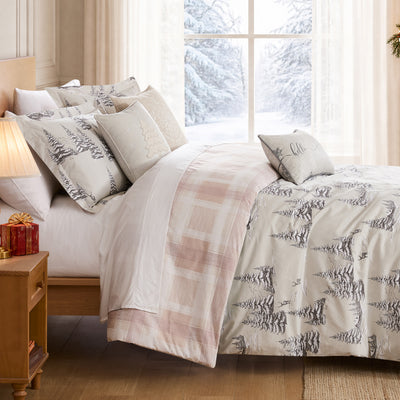 Christmas comforter set on a bed with a cream Frosted Pines print of gray snowy pine trees and deer, blush plaid reverse folded back, coordinating pine shams, snowflake and Christmas tree throw pillows, and a hello winter lumbar pillow, styled beside a lamp-lit nightstand with red wrapped gifts and a snowy window view.