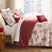 Winterwood Christmas comforter set styled on a bed with a cream ground red toile print of deer, pine trees, snowmen, and cabins, red windowpane plaid reverse folded back, matching print shams, a cream Joy pillow, a red and white peppermint swirl round pillow, and a red snowflake lumbar pillow, beside a lamp-lit nightstand with red gifts and a snowy window view.