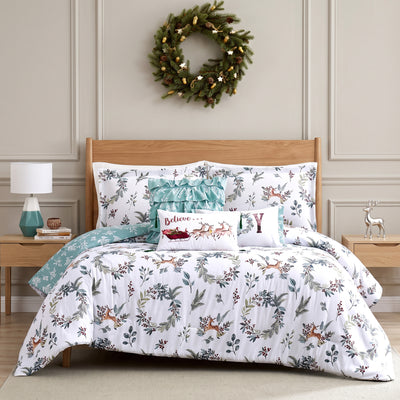 Happy Holidays comforter set styled on a bed with a cream upholstered headboard against a wood plank wall, white comforter with watercolor greenery and leaping deer print and sage-green reverse, matching shams, a sage ruffled pillow, a Believe Santa-sleigh lumbar pillow, and a JOY wreath lumbar pillow, beside a glass lamp and wood nightstand, under a pine wreath, with a lit wood stove and a decorated Christmas tree.