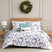 Happy Holidays comforter set styled on a bed with a cream upholstered headboard against a wood plank wall, white comforter with watercolor greenery and leaping deer print and sage-green reverse, matching shams, a sage ruffled pillow, a Believe Santa-sleigh lumbar pillow, and a JOY wreath lumbar pillow, beside a glass lamp and wood nightstand, under a pine wreath, with a lit wood stove and a decorated Christmas tree.