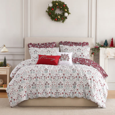 Holiday Harmony comforter set styled on a bed with a cream upholstered headboard against a paneled wall, white comforter with a red and sage folk-floral damask print and a burgundy winter-forest reverse, matching shams, burgundy ruffled Euro shams, a red Merry lumbar pillow, and a white floral poinsettia throw pillow, beside a white nightstand and lamp, under an ornament wreath.