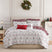 Holiday Harmony comforter set styled on a bed with a cream upholstered headboard against a paneled wall, white comforter with a red and sage folk-floral damask print and a burgundy winter-forest reverse, matching shams, burgundy ruffled Euro shams, a red Merry lumbar pillow, and a white floral poinsettia throw pillow, beside a white nightstand and lamp, under an ornament wreath.