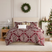 Christmas Carol comforter set styled on a bed with a cream upholstered headboard against a paneled wall, burgundy comforter with cream floral damask print and red plaid reverse, matching shams, a faux fur pillow, and a cream Merry lumbar pillow, beside a white lamp and wood nightstand, under a pine wreath, with a decorated Christmas tree and a white mantel.