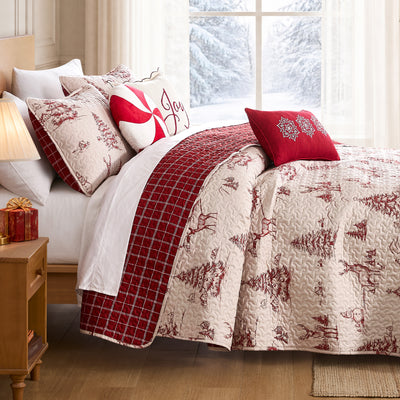 Winterwood Christmas quilt set styled on a bed with a cream ground red toile winter-scene print of deer, pine trees, snowmen, and cabins, machine-quilted, red windowpane plaid reverse folded back, coordinating quilted shams, a cream Joy pillow, a red and white peppermint swirl round pillow, and a red snowflake lumbar pillow, beside a lamp-lit nightstand with red gifts and a snowy window view.
