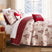 Winterwood Christmas quilt set styled on a bed with a cream ground red toile winter-scene print of deer, pine trees, snowmen, and cabins, machine-quilted, red windowpane plaid reverse folded back, coordinating quilted shams, a cream Joy pillow, a red and white peppermint swirl round pillow, and a red snowflake lumbar pillow, beside a lamp-lit nightstand with red gifts and a snowy window view.