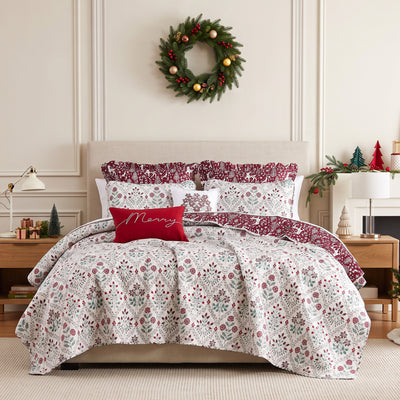 Holiday Harmony quilt set styled on a bed with a cream upholstered headboard against a paneled wall, white machine-quilted red and sage folk-floral damask print with a burgundy winter-forest reverse, matching shams, burgundy ruffled Euro shams, a red Merry lumbar pillow, and a white floral poinsettia throw pillow, beside a white lamp and wood nightstand, under an ornament wreath.