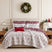 Holiday Harmony quilt set styled on a bed with a cream upholstered headboard against a paneled wall, white machine-quilted red and sage folk-floral damask print with a burgundy winter-forest reverse, matching shams, burgundy ruffled Euro shams, a red Merry lumbar pillow, and a white floral poinsettia throw pillow, beside a white lamp and wood nightstand, under an ornament wreath.