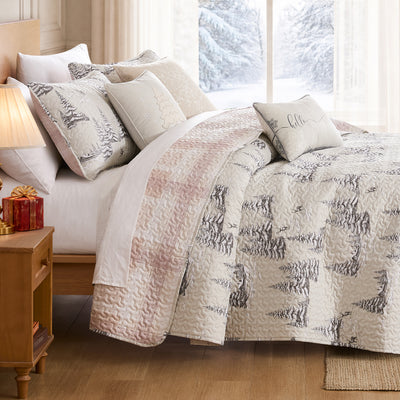 Frosted Pines Christmas quilt set styled on a bed with a cream and gray machine-quilted winter forest print of snowy pine trees and deer, soft blush plaid reverse folded back, coordinating quilted shams, embroidered snowflake and Christmas tree throw pillows, and a hello winter lumbar pillow, beside a lamp-lit nightstand with red gifts and a snowy window view.