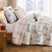 Frosted Pines Christmas quilt set styled on a bed with a cream and gray machine-quilted winter forest print of snowy pine trees and deer, soft blush plaid reverse folded back, coordinating quilted shams, embroidered snowflake and Christmas tree throw pillows, and a hello winter lumbar pillow, beside a lamp-lit nightstand with red gifts and a snowy window view.