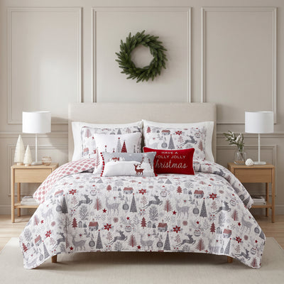 Holly Jolly Lane quilt set styled on a bed with a cream upholstered headboard against a paneled wall, white ground gray and red winter print quilt with red snowflake reverse, matching shams, a Christmas trees pillow, a reindeer pom-pom lumbar pillow, and a red Have a Holly Jolly Christmas lumbar pillow, flanked by wood nightstands with white lamps, under a pine wreath.