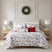 Holly Jolly Lane quilt set styled on a bed with a cream upholstered headboard against a paneled wall, white ground gray and red winter print quilt with red snowflake reverse, matching shams, a Christmas trees pillow, a reindeer pom-pom lumbar pillow, and a red Have a Holly Jolly Christmas lumbar pillow, flanked by wood nightstands with white lamps, under a pine wreath.