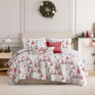 Merry and Bright quilt set styled on a bed with a cream upholstered headboard against a paneled wall, white machine-quilted red and gray geometric Christmas trees with red stars, matching shams, a red Merry and Bright pom-pom throw pillow, a white Christmas tree pom-pom pillow, and a festive tree lumbar pillow, beside a white lamp and nightstand, under an ornament wreath.