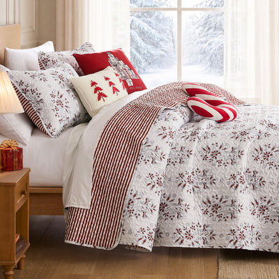Merry Manor Christmas quilt set styled on a bed with a white ground red and gray machine-quilted floral, poinsettia, and reindeer print, red ticking stripe reverse folded back, coordinating quilted shams, a red Happy Holidays gift-tree pillow, a cream red-tree lumbar pillow, and a candy cane bolster, beside a lamp-lit nightstand with red gifts and a snowy window view.