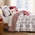 Merry Manor Christmas quilt set styled on a bed with a white ground red and gray machine-quilted floral, poinsettia, and reindeer print, red ticking stripe reverse folded back, coordinating quilted shams, a red Happy Holidays gift-tree pillow, a cream red-tree lumbar pillow, and a candy cane bolster, beside a lamp-lit nightstand with red gifts and a snowy window view.