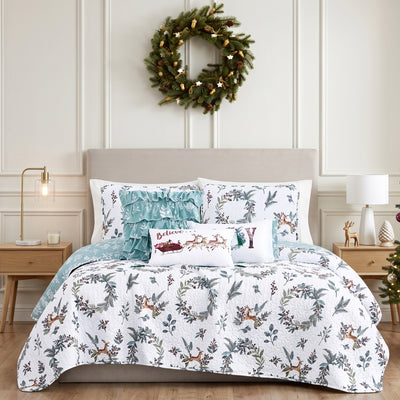 Happy Holidays quilt set styled on a bed with a cream upholstered headboard against a paneled wall, white machine-quilted greenery, berries, and leaping deer print with sage-green reverse, matching shams, a sage ruffled pillow, a Believe Santa-sleigh-and-reindeer lumbar pillow, and a JOY wreath lumbar pillow, beside wood nightstands with brass and white lamps, under a pine wreath, with a decorated Christmas tree.