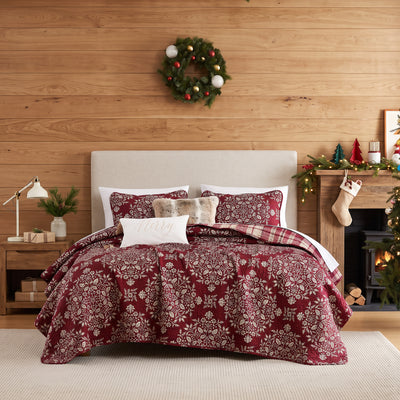 Christmas Carol quilt set styled on a bed against a wood plank wall, burgundy quilt with cream floral damask print and red plaid reverse, matching shams, a faux fur lumbar pillow, and a cream Merry lumbar pillow, beside a white lamp and wood nightstand, under an ornament wreath, with a lit Christmas tree and a wood stove.