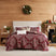 Christmas Carol quilt set styled on a bed against a wood plank wall, burgundy quilt with cream floral damask print and red plaid reverse, matching shams, a faux fur lumbar pillow, and a cream Merry lumbar pillow, beside a white lamp and wood nightstand, under an ornament wreath, with a lit Christmas tree and a wood stove.