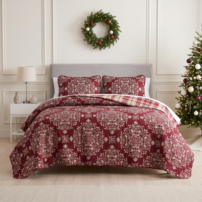 Christmas Carol oversized quilt set styled on a bed with a gray upholstered headboard, burgundy quilt with cream floral damask print and red and cream plaid reverse, matching shams, beside a white nightstand with a glass lamp, under an ornament wreath, with a decorated Christmas tree.