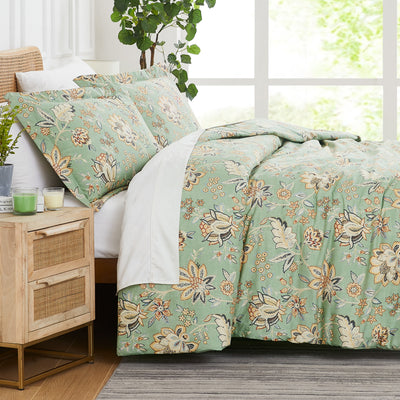 Side view of the Jacobean Willow Green comforter and matching shams on a bed with white sheets, a rattan nightstand and a leafy plant #color_jacobean-willow-green_15439115124962