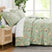 Side view of the Jacobean Willow Green comforter and matching shams on a bed with white sheets, a rattan nightstand and a leafy plant #color_jacobean-willow-green_15439115124962