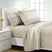 A neatly made bed with beige and off-white striped bedding, including two pillows and a matching duvet cover, shown in a bright bedroom setting with a white nightstand and window. #color_quiet-horizon-taupe