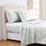 A neatly made bed with a white sheet set and matching pillowcases featuring a delicate green floral pattern, styled in a bright bedroom with a cream tufted headboard and window curtains. #color_shamrock-breeze-light-gray