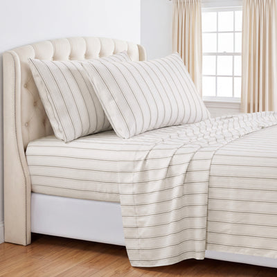 A neatly made bed with a cream-colored tufted headboard, matching striped pillows, and a cream sheet set with thin gray stripes, set in a bright bedroom with beige curtains and a window. #color_stone-harbor-taupe