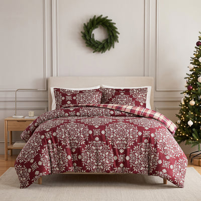 Christmas Carol oversized comforter set styled on a bed with a cream upholstered headboard against a paneled wall, burgundy comforter with cream floral damask print and red and cream plaid reverse folded back, matching shams, beside a wood nightstand, with a decorated Christmas tree.