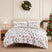 Holly Jolly Lane oversized comforter set styled on a bed with a cream upholstered headboard against a paneled wall, white ground gray and red winter print comforter with red snowflake reverse and matching shams, beside a brass lamp and wood nightstand on a jute rug, under a pine wreath, with a decorated Christmas tree.