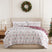 Holiday Harmony oversized comforter set styled on a bed with a cream upholstered headboard against a paneled wall, white comforter with a red and sage folk-floral damask print and a burgundy winter-forest reverse, matching shams, beside a brass lamp and wood nightstand, under an ornament wreath, with a decorated Christmas tree.