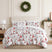 Merry and Bright oversized comforter set styled on a bed with a cream upholstered headboard against a paneled wall, white comforter with red and gray geometric Christmas trees and red stars, matching shams, beside a white lamp and wood nightstand, under a pinecone wreath.