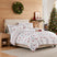Holly Jolly Lane Duvet Cover Set styled on a bed with a cream upholstered headboard, white ground gray and red Scandinavian winter print of deer, pine trees, cottages, ornaments, and poinsettias with red snowflake reverse and matching shams, beside a wood nightstand with a brass lamp, under an ornament wreath, with a lit Christmas tree with red bows.