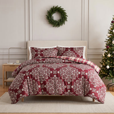 Christmas Carol duvet cover set styled on a bed with a cream upholstered headboard against a paneled wall, burgundy duvet cover with cream floral damask print and red and cream plaid reverse folded back, matching shams, beside a wood nightstand, with a decorated Christmas tree.