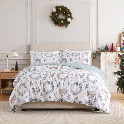 Happy Holidays duvet cover set styled on a bed with a cream upholstered headboard against a paneled wall, white duvet cover with a watercolor print of winter greenery, berries, and leaping deer and a sage-green botanical reverse folded back, matching shams, beside a wood nightstand with a brass lamp, under an ornament wreath, with a decorated Christmas tree.