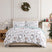 Happy Holidays duvet cover set styled on a bed with a cream upholstered headboard against a paneled wall, white duvet cover with a watercolor print of winter greenery, berries, and leaping deer and a sage-green botanical reverse folded back, matching shams, beside a wood nightstand with a brass lamp, under an ornament wreath, with a decorated Christmas tree.