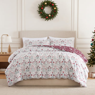 Holiday Harmony duvet cover set styled on a bed with a cream upholstered headboard against a paneled wall, white duvet cover with a red and sage folk-floral damask print and a burgundy winter-forest reverse, matching shams, beside a brass lamp and wood nightstand, under an ornament wreath, with a decorated Christmas tree.