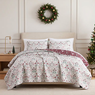 Holiday Harmony oversized quilt set styled on a bed with a cream upholstered headboard against a paneled wall, white machine-quilted red and sage folk-floral damask print with a burgundy winter-forest reverse, matching shams, beside a brass lamp and wood nightstand, under an ornament wreath, with a decorated Christmas tree.