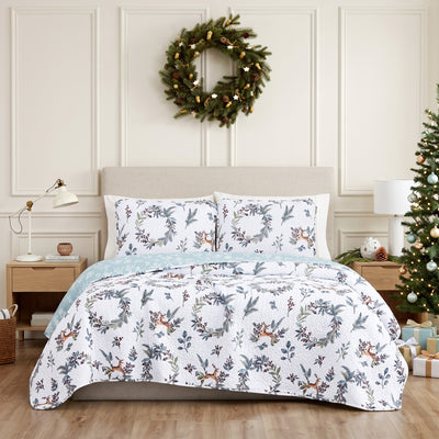 Happy Holidays quilt set styled on a bed with a cream upholstered headboard against a paneled wall, white machine-quilted greenery, berries, and leaping deer print with sage-green reverse folded back, matching shams, beside a wood nightstand with a brass lamp, under an ornament wreath, with a decorated Christmas tree and wrapped gifts.