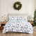 Happy Holidays quilt set styled on a bed with a cream upholstered headboard against a paneled wall, white machine-quilted greenery, berries, and leaping deer print with sage-green reverse folded back, matching shams, beside a wood nightstand with a brass lamp, under an ornament wreath, with a decorated Christmas tree and wrapped gifts.